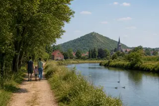 A family walks along a countryside path in the Scarpe-Escaut Regional Natural Park, surrounded by meadows, trees, a canal, and distant houses with old mining heaps covered in greenery.