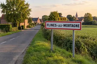 Panneau d’entrée Flines-lez-Mortagne au bord d’une route de campagne, maisons en brique et arbres sous un ciel clair du nord de la France.